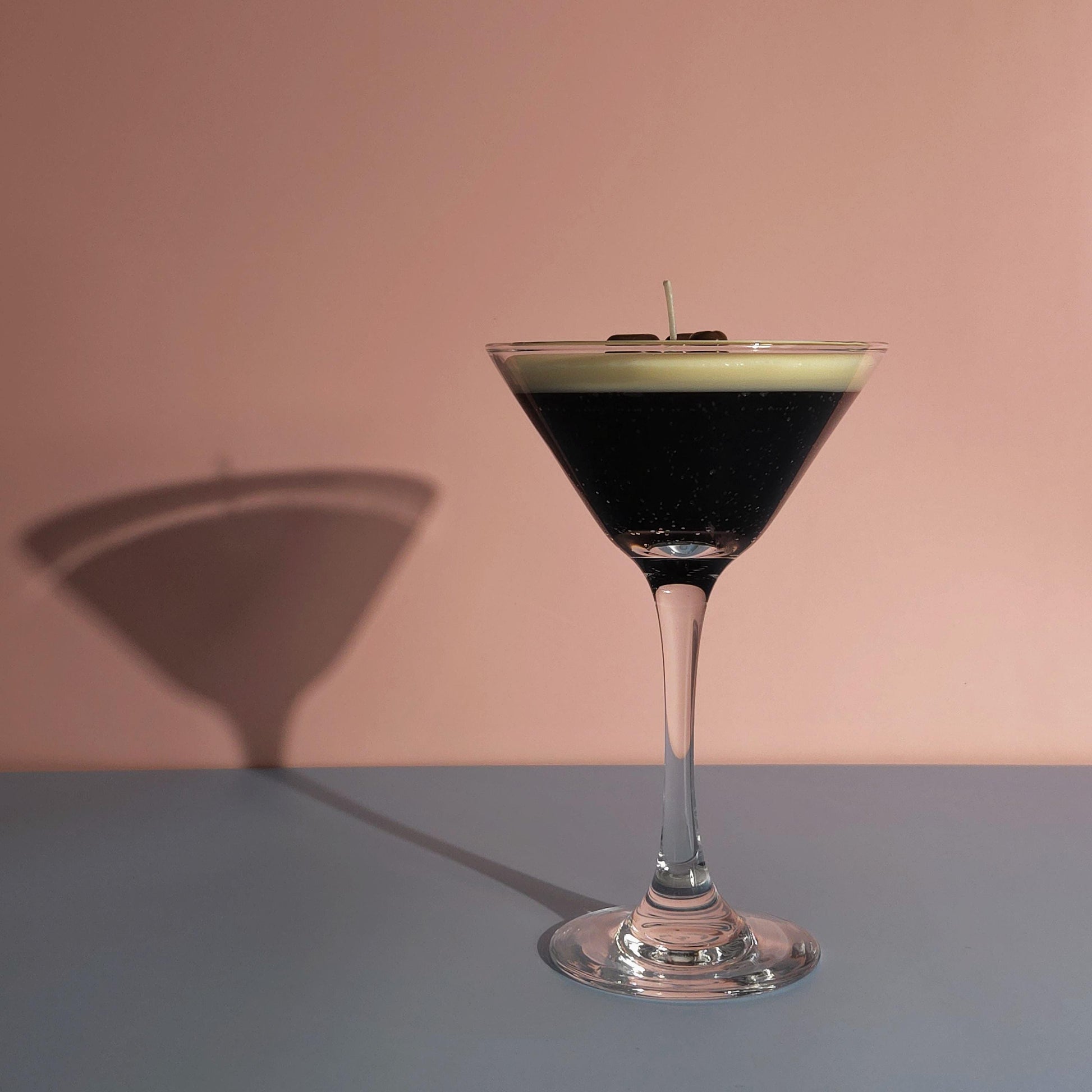 A realistic espresso martini candle in a classic martini glass, featuring a creamy frothy wax top with three coffee bean garnishes. The candle sits on casting a soft shadow against a pastel pink and blue background. Hand-poured soy wax candle, coffee scented candle, candle gifts for coffee lover, cocktail candle, drink lover gifts, birthday gifts, candle with container, handmade candle, coffee beans