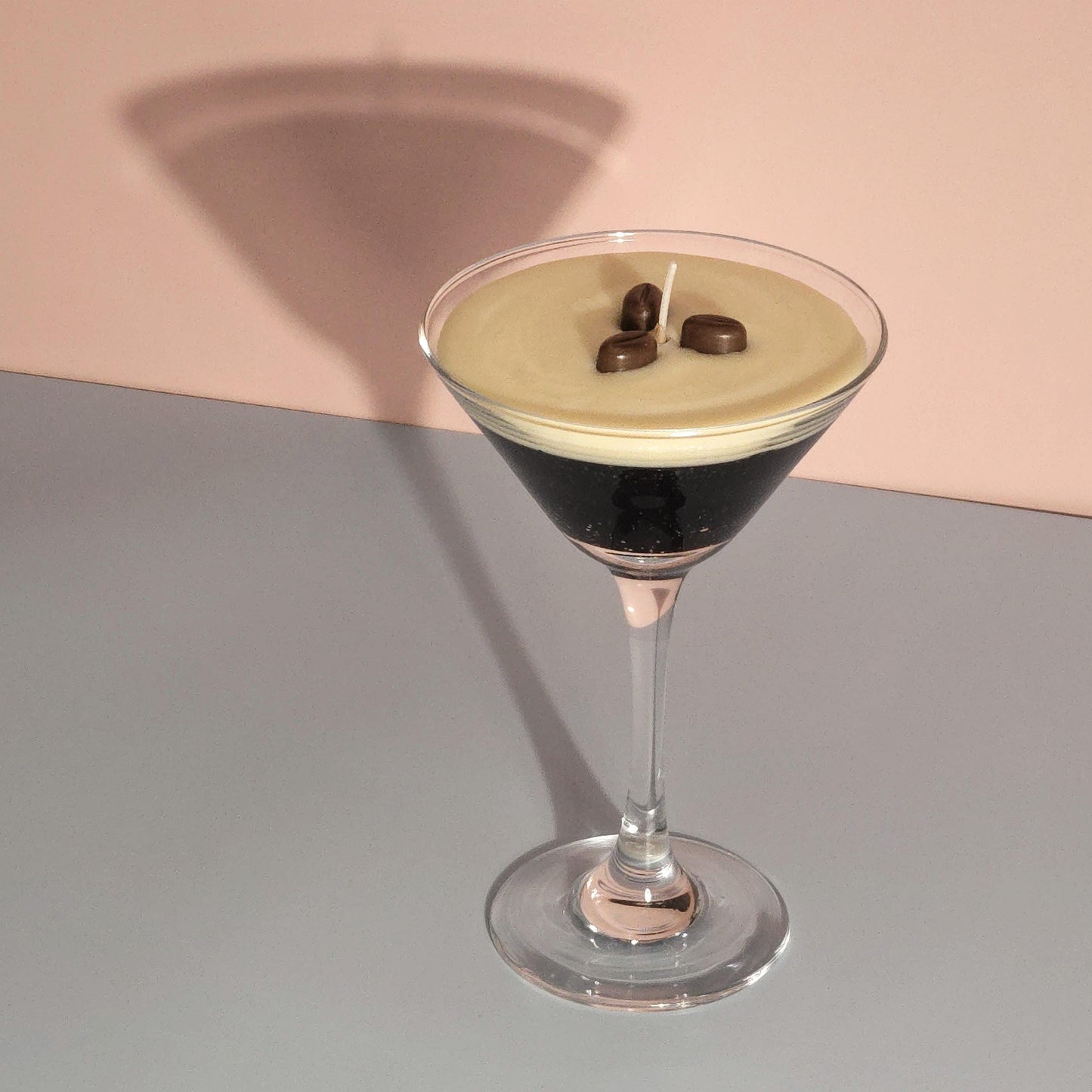 A realistic espresso martini candle in a classic martini glass, featuring a creamy frothy wax top with three coffee bean garnishes. The candle sits on casting a soft shadow against a pastel pink and blue background. Hand-poured soy wax candle, coffee scented candle, candle gifts for coffee lover, cocktail candle, drink lover gifts, birthday gifts, candle with container, handmade candle, coffee beans