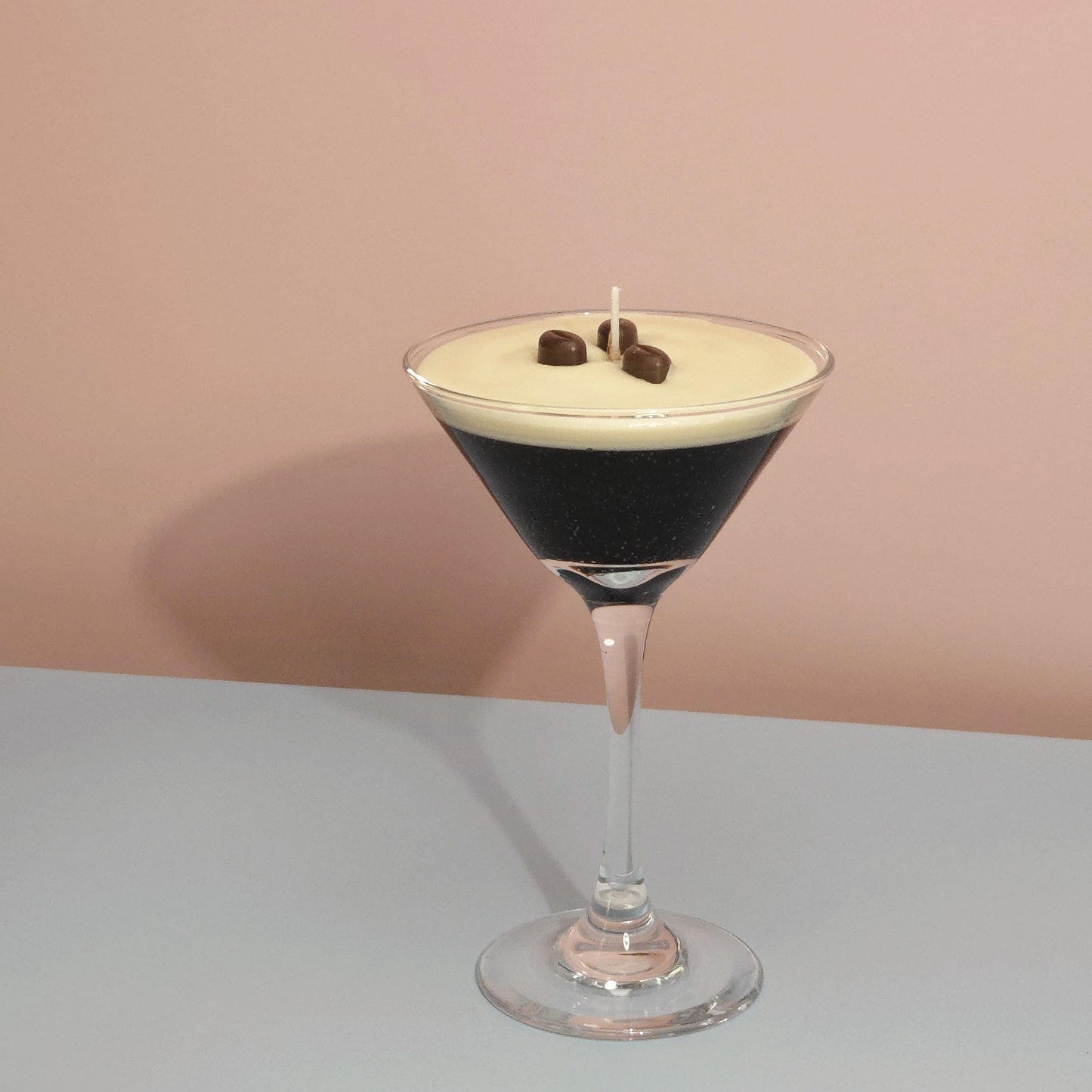 A realistic espresso martini candle in a classic martini glass, featuring a creamy frothy wax top with three coffee bean garnishes. The candle sits on casting a soft shadow against a pastel pink and blue background. Hand-poured soy wax candle, coffee scented candle, candle gifts for coffee lover, cocktail candle, drink lover gifts, birthday gifts, candle with container, handmade candle, coffee beans