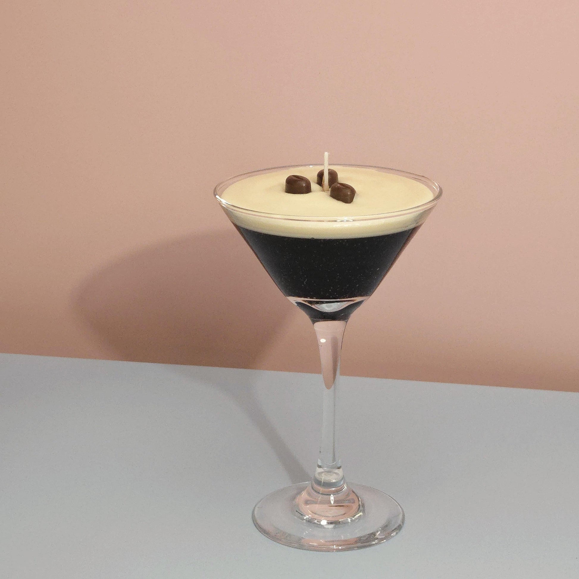 A realistic espresso martini candle in a classic martini glass, featuring a creamy frothy wax top with three coffee bean garnishes. The candle sits on casting a soft shadow against a pastel pink and blue background. Hand-poured soy wax candle, coffee scented candle, candle gifts for coffee lover, cocktail candle, drink lover gifts, birthday gifts, candle with container, handmade candle, coffee beans