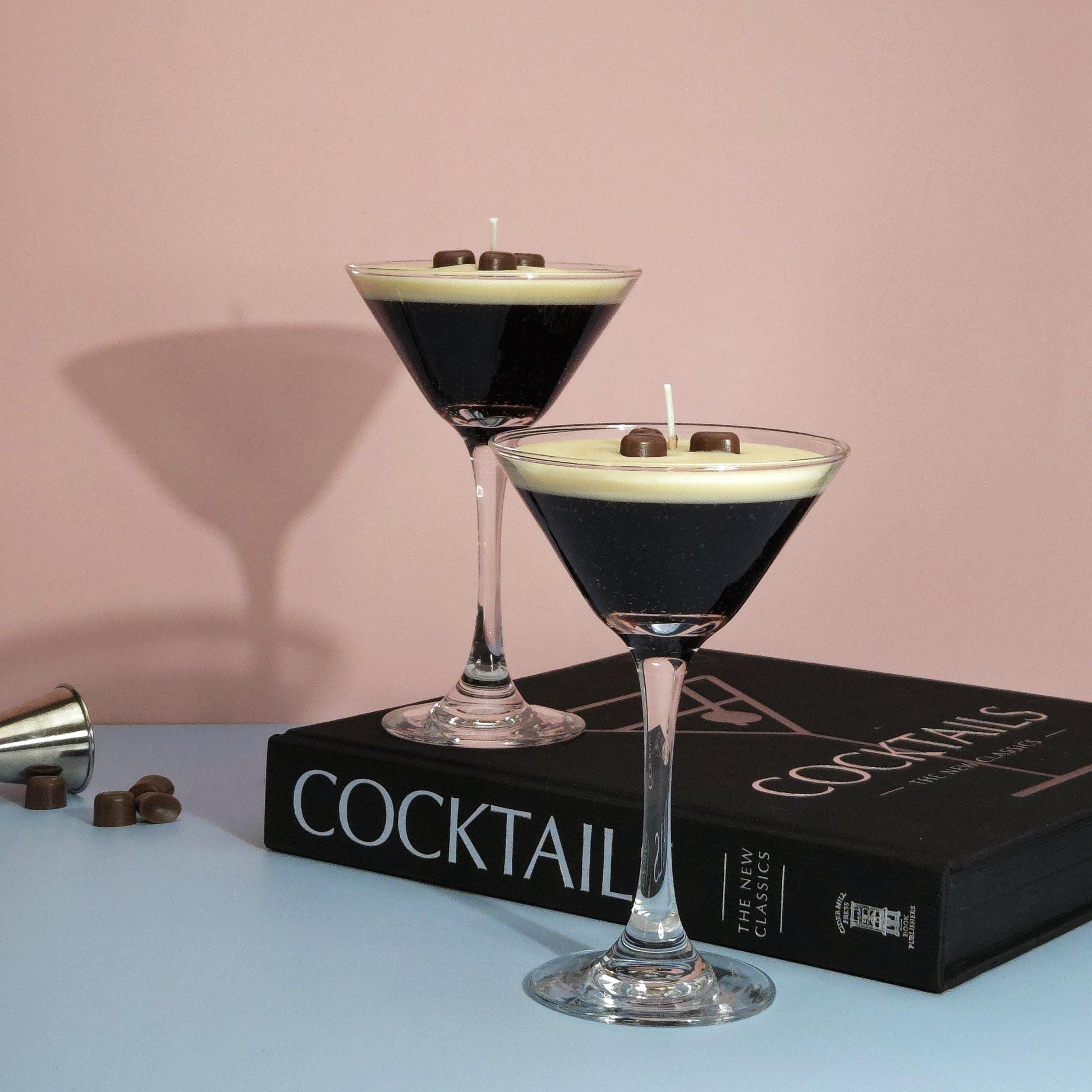 A realistic espresso martini candle in a classic martini glass, featuring a creamy frothy wax top with three coffee bean garnishes. The candle sits on a cocktail book, casting a soft shadow against a pastel pink and blue background. Hand-poured soy wax candle, coffee scented candle, candle gifts for coffee lover, cocktail candle, drink lover gifts, birthday gifts, candle with container, handmade candle, coffee beans