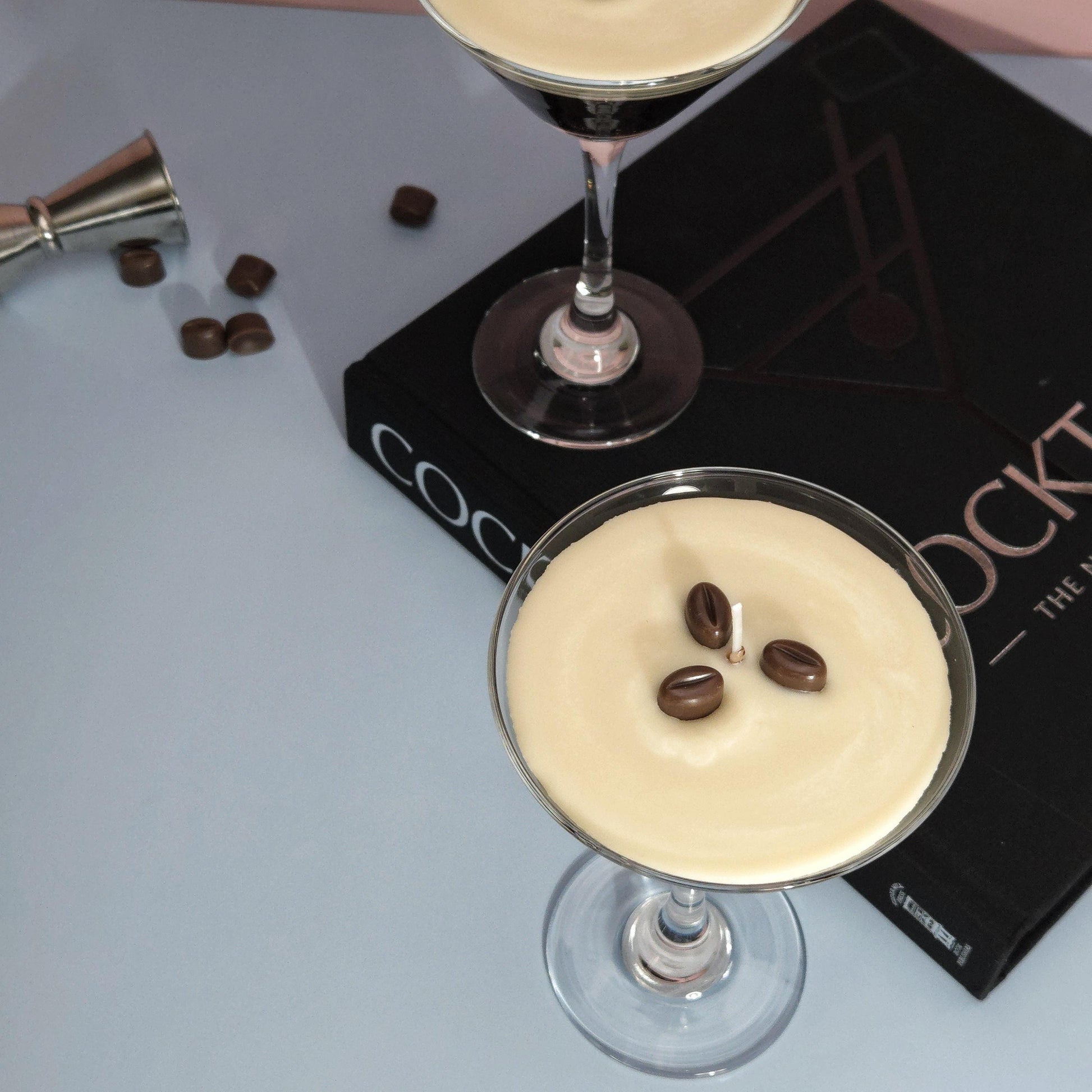A realistic espresso martini candle in a classic martini glass, featuring a creamy frothy wax top with three coffee bean garnishes. The candle sits on a cocktail book, casting a soft shadow against a pastel pink and blue background. Hand-poured soy wax candle, coffee scented candle, candle gifts for coffee lover, cocktail candle, drink lover gifts, birthday gifts, candle with container, handmade candle, coffee beans