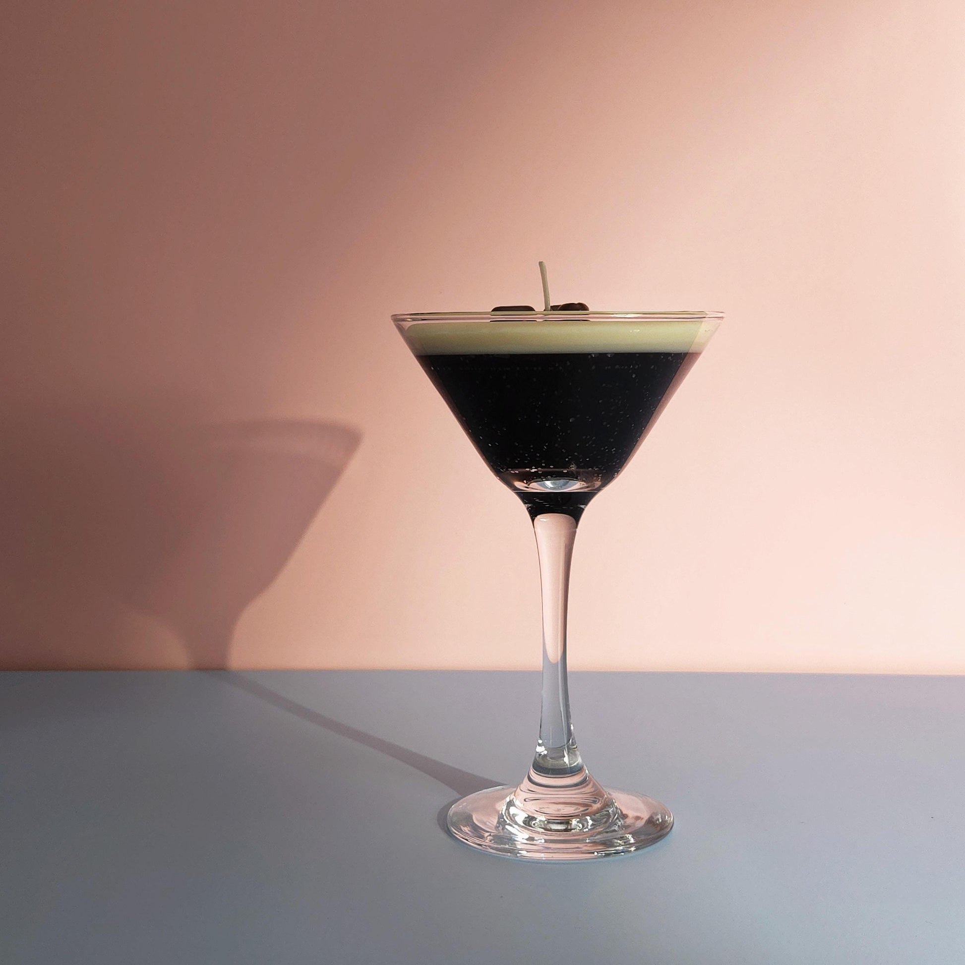 A realistic espresso martini candle in a classic martini glass, featuring a creamy frothy wax top with three coffee bean garnishes. The candle sits on casting a soft shadow against a pastel pink and blue background. Hand-poured soy wax candle, coffee scented candle, candle gifts for coffee lover, cocktail candle, drink lover gifts, birthday gifts, candle with container, handmade candle, coffee beans