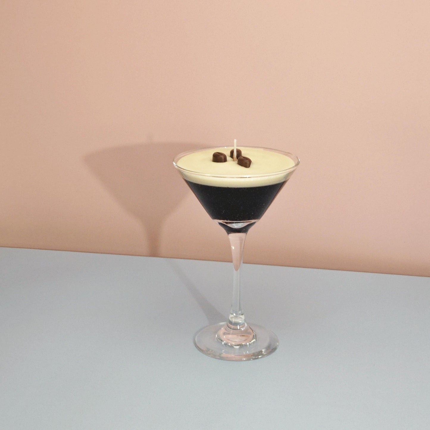 A realistic espresso martini candle in a classic martini glass, featuring a creamy frothy wax top with three coffee bean garnishes. The candle sits on casting a soft shadow against a pastel pink and blue background. Hand-poured soy wax candle, coffee scented candle, candle gifts for coffee lover, cocktail candle, drink lover gifts, birthday gifts, candle with container, handmade candle, coffee beans