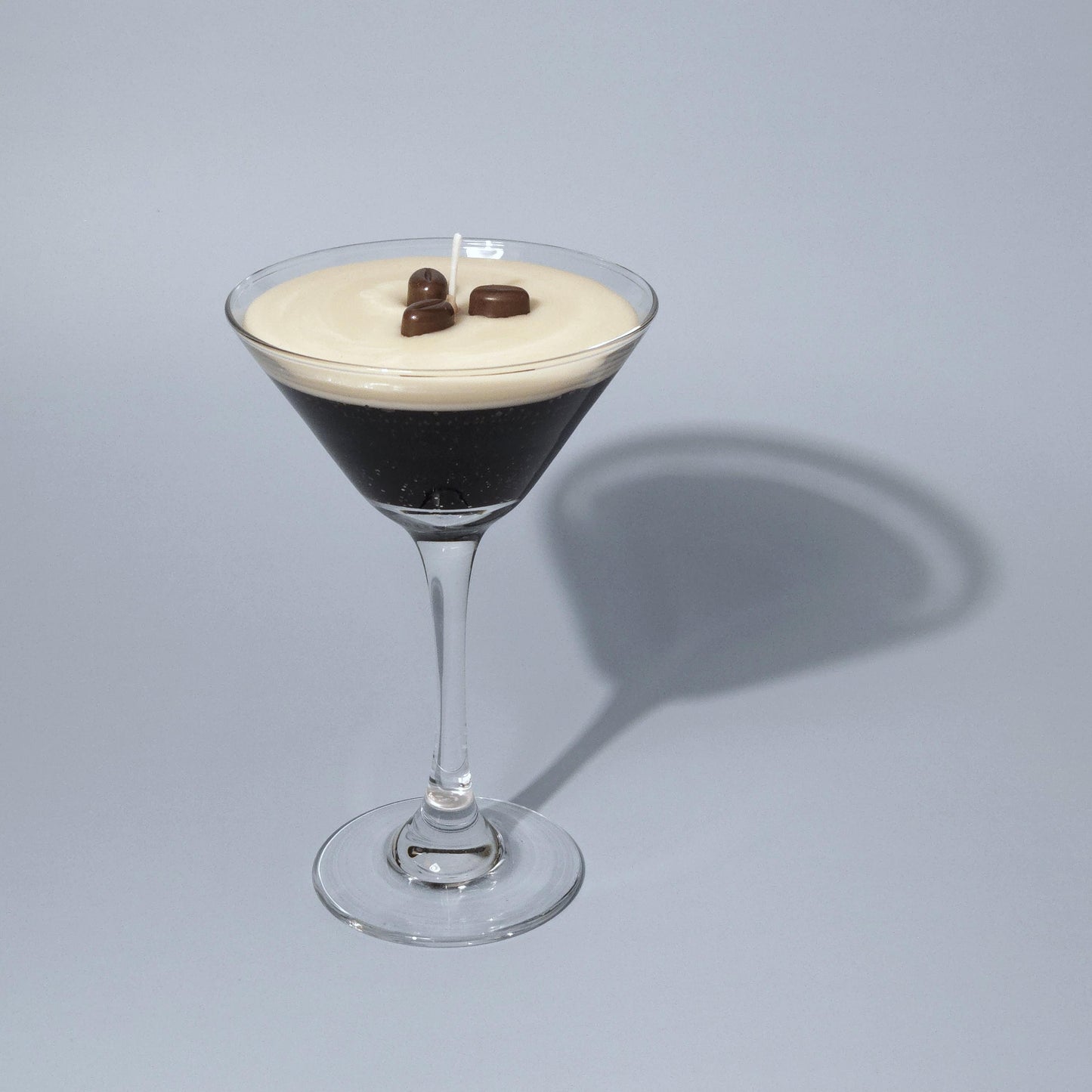 A realistic espresso martini candle in a classic martini glass, featuring a creamy frothy wax top with three coffee bean garnishes. The candle sits on casting a soft shadow against a pastel blue background. Hand-poured soy wax candle, coffee scented candle, candle gifts for coffee lover, cocktail candle, drink lover gifts, birthday gifts, candle with container, handmade candle, coffee beans