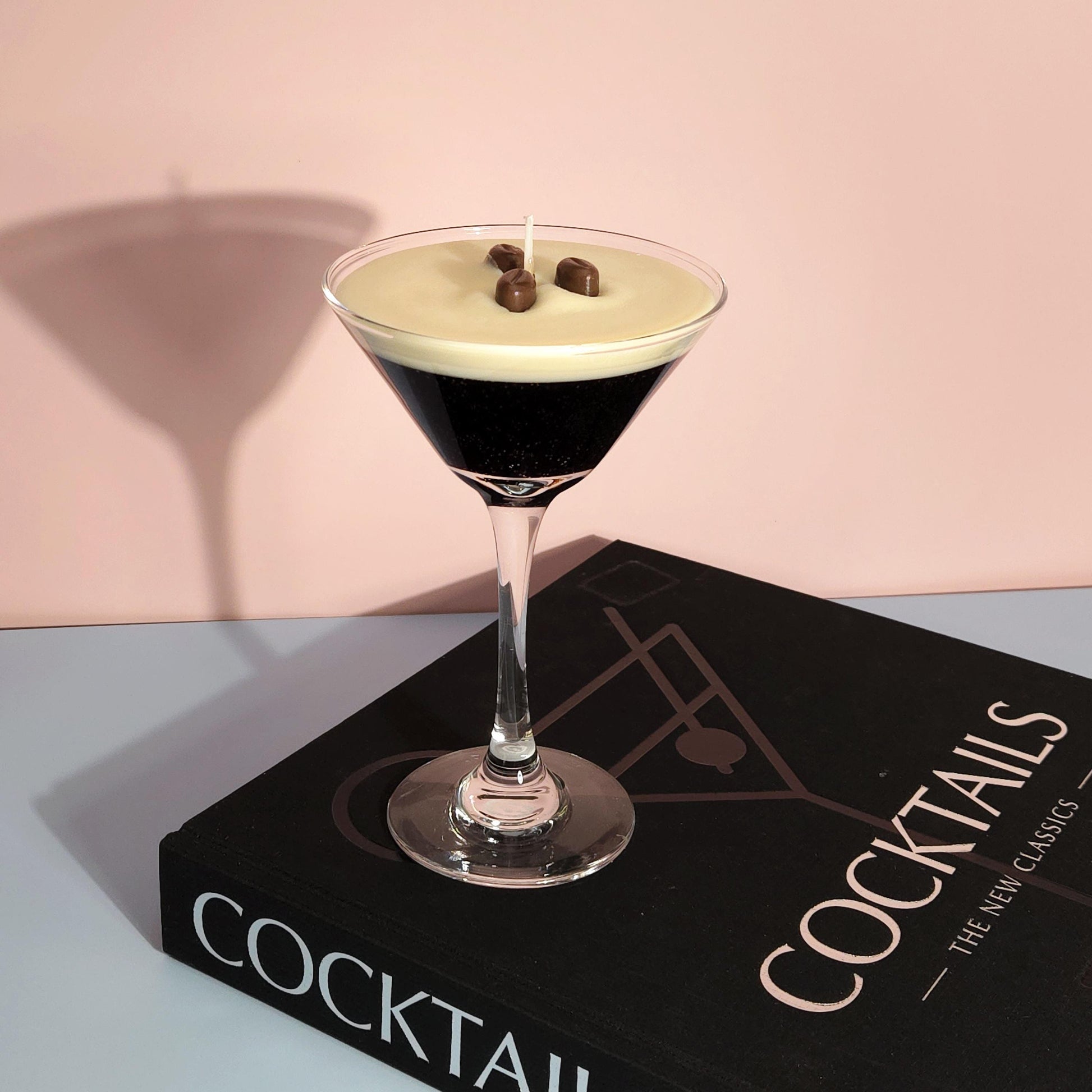 A realistic espresso martini candle in a classic martini glass, featuring a creamy frothy wax top with three coffee bean garnishes. The candle sits on a cocktail book, casting a soft shadow against a pastel pink and blue background. Hand-poured soy wax candle, coffee scented candle, candle gifts for coffee lover, cocktail candle, drink lover gifts, birthday gifts, candle with container, handmade candle, coffee beans