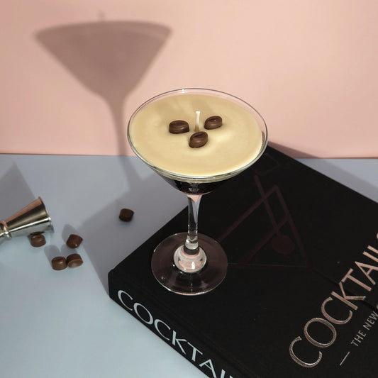 A realistic espresso martini candle in a classic martini glass, featuring a creamy frothy wax top with three coffee bean garnishes. The candle sits on a cocktail book, casting a soft shadow against a pastel pink and blue background. Hand-poured soy wax candle, coffee scented candle, candle gifts for coffee lover, cocktail candle, drink lover gifts, birthday gifts, candle with container, handmade candle, coffee beans